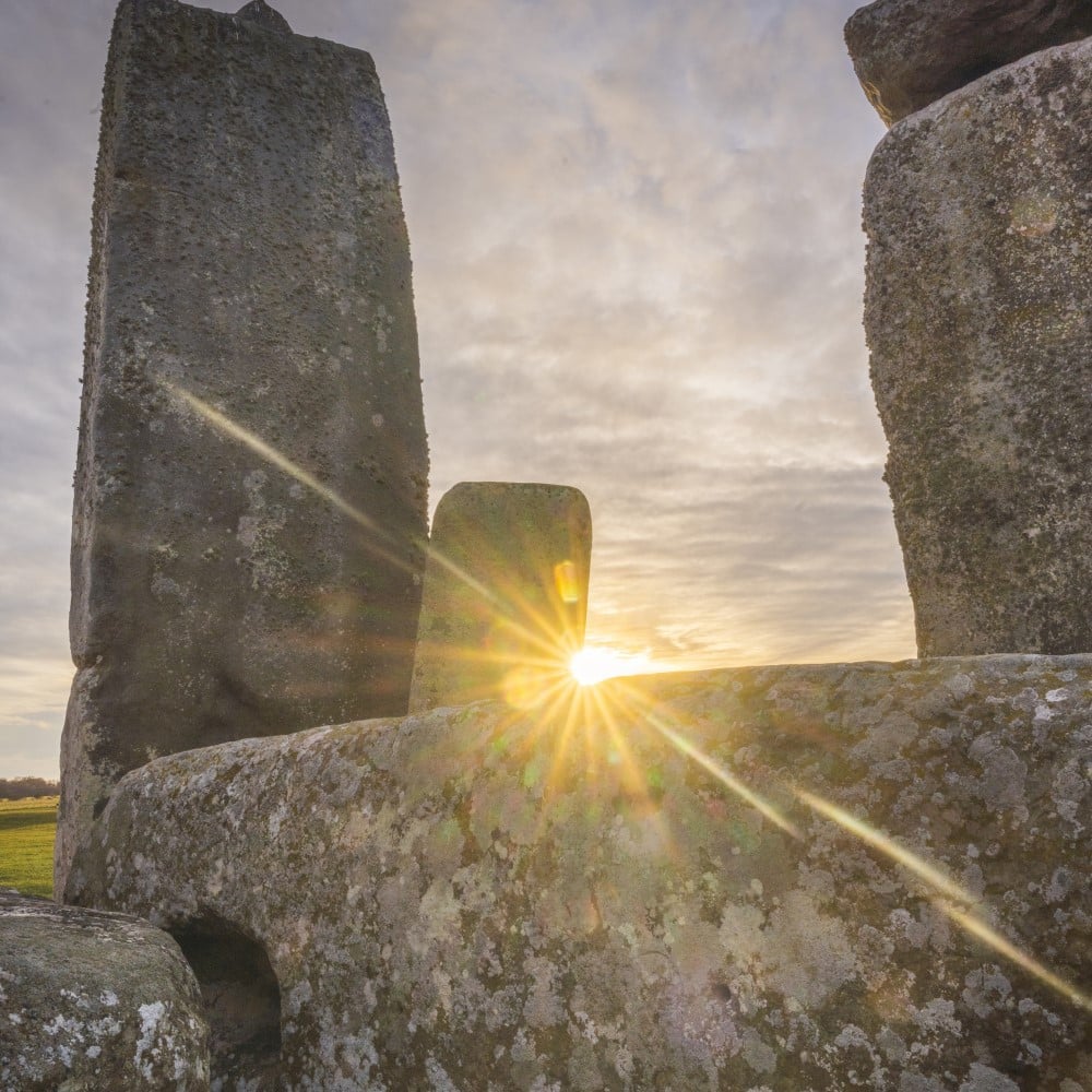 Photo of the sun shining low in the sky through Stonehenge