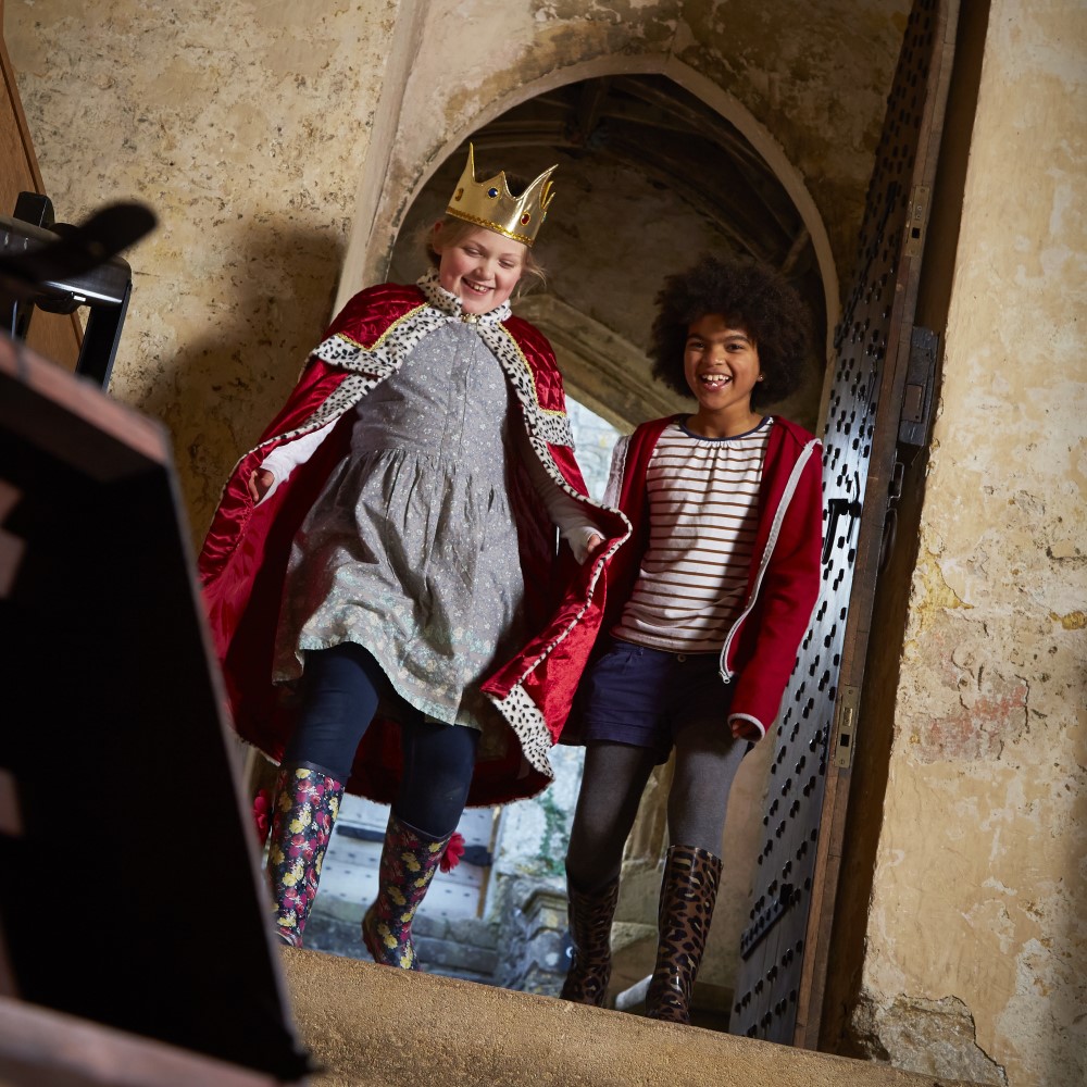 Photo of two children, one dressed as a monarch, laughing and walking through a doorway at Farleigh Hungerford Castle