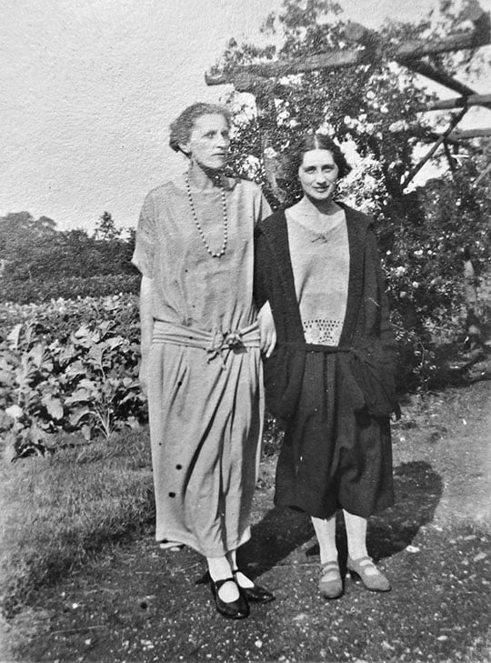 Black and white photograph of Winifred Holtby (tall and blonde) and Vera Brittain (shorter with dark hair) standing outside with arms linked