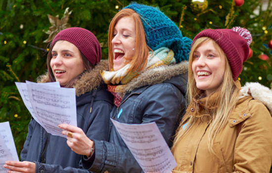 Three young women singing Christmas carols outdoors in front of a decorated Christmas tree. They are dressed warmly in coats, scarves, and hats, holding sheet music and smiling as they sing. Three young women singing Christmas carols outdoors in front of a decorated Christmas tree. They are dressed warmly in coats, scarves, and hats, holding sheet music and smiling as they sing.